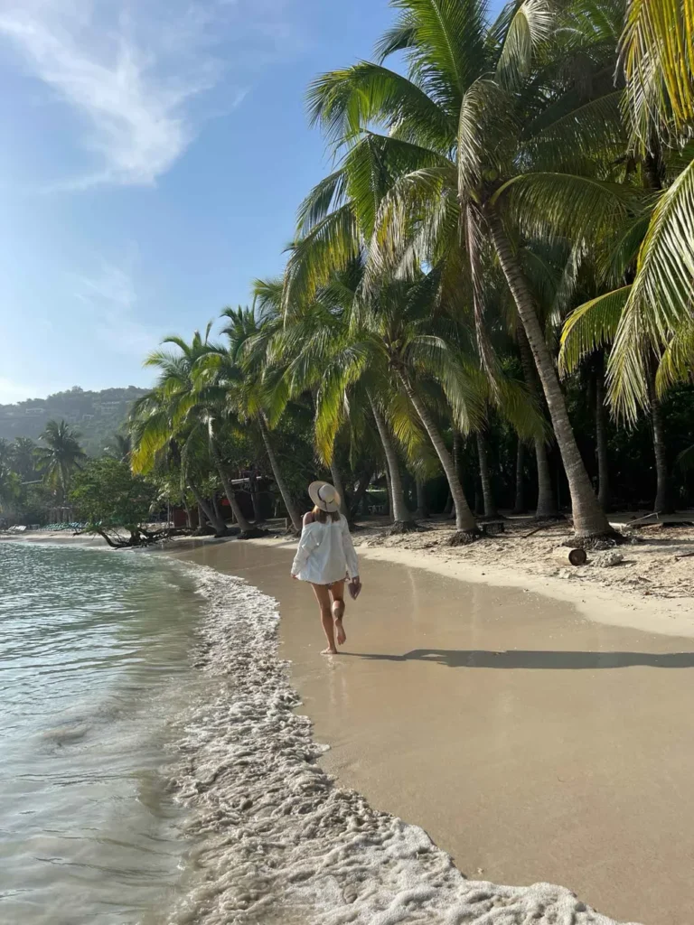 private beach at Casa Las Gatas in Zihuatanejo – truly one of the best unique places to stay in Mexico woman in a white shirt with a sunhat holding her sandals walking on a white sand beach with light waves next to a lot of palm trees in mexican nature at casa las gatas, a perfect unique stay in mexico cose to Zihuatanejo