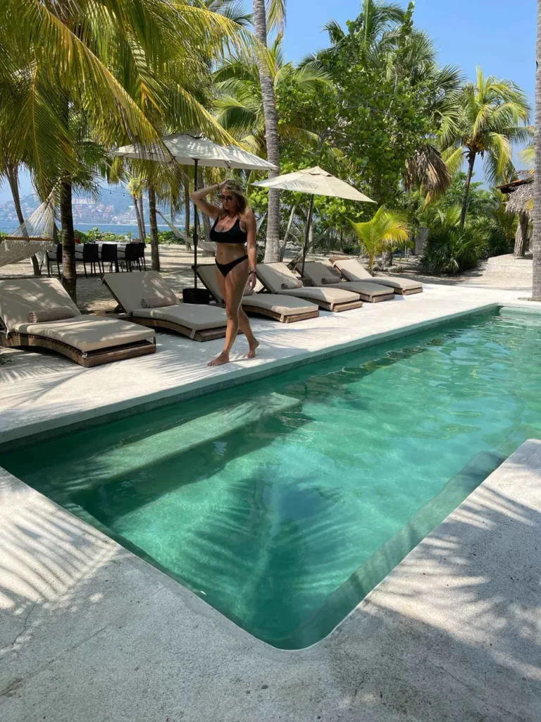 private pool vibes a blonde woman in a black bikini walking next to a long rectangular turquoise pool surrounded by laychairs sun umbrellas and palm trees at casa las gatas in Mexico close to Zihuatanejo