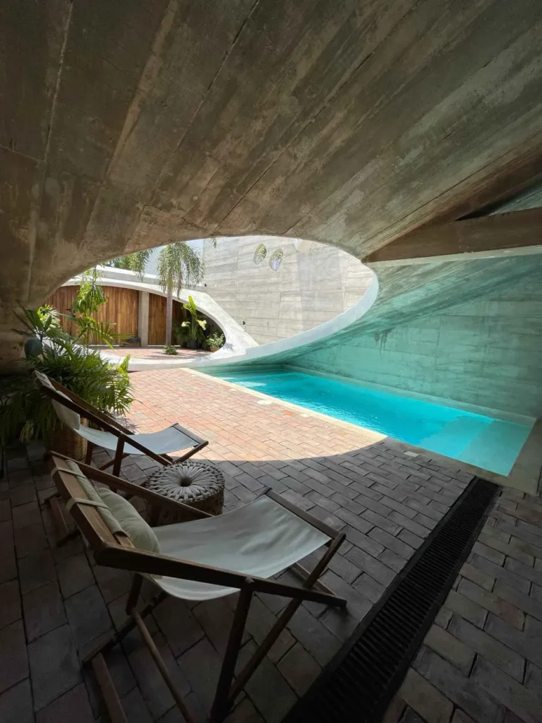 Casa VO in Puerto Escondido two modern laychairs and a stool standing in front of a turquoise pool and plants inside modern architecture with a diagonal roof and a round hole letting the sun light inthe back is a partially wooden and stone area