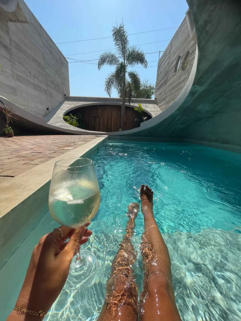 one of my favorite unique stays in mexico woman laying in a turquoise clear pool holding a glass of white vine overlooking a modern archticture in grey with a roof with an angle and hole seeing anoter hole and a plam tree in the back at casa von in puerto escondido