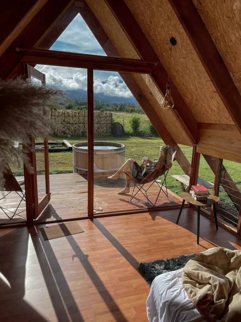 A-frame cabin with a view. Etlan Cabanas photo from inside the A-frame cabin Etlan Cabanas in Mexico with a wooden roof a glass front and a jacuzzi outside surrounded by mexican nature and a woman sitting outside in a chair
