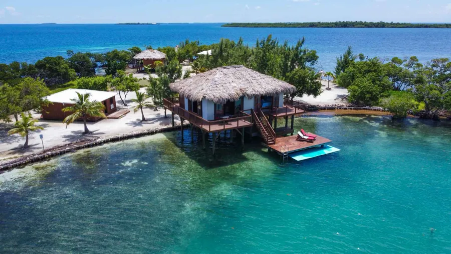 small island with greenery and some housing including overwater bungalow surrounded by blue clear water and small blue hole, one of the best hidden gems in Belize: private island rental in Belize - Little Peter Oasis