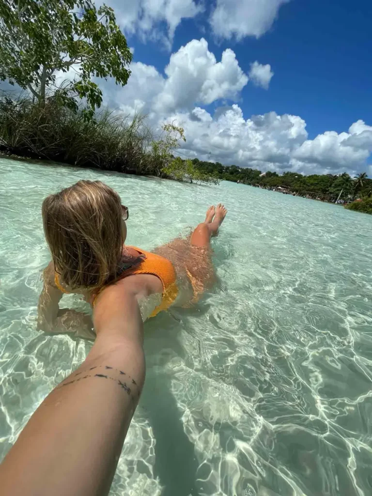 blonde woman taking a picture of herself from the back laying in clear water with some forest or jungle landscape around at the Cenote Cocalitos Beach Resort in Bacalar Mexico 