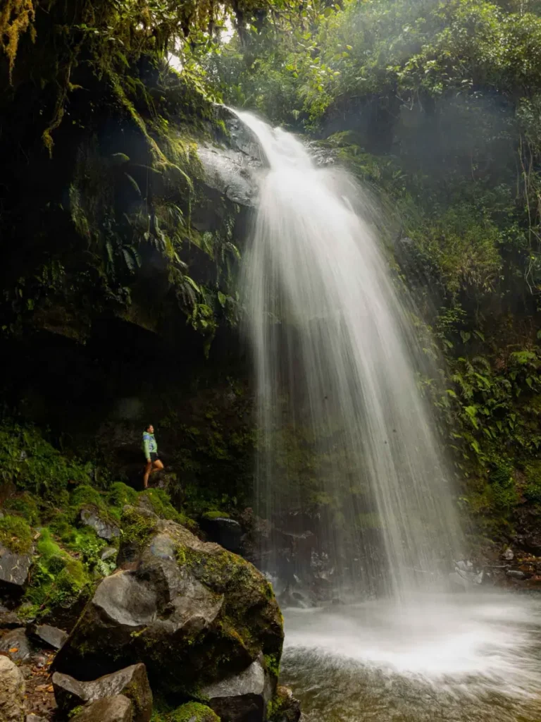 woman with a green rainjacket standing behind a large waterfall pouring from a rock cave in the jungle