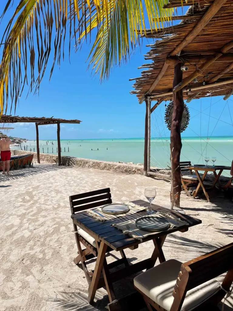 beach hut with seating are with table and chairs with plated table and more wooden huts on a white beach with clear water