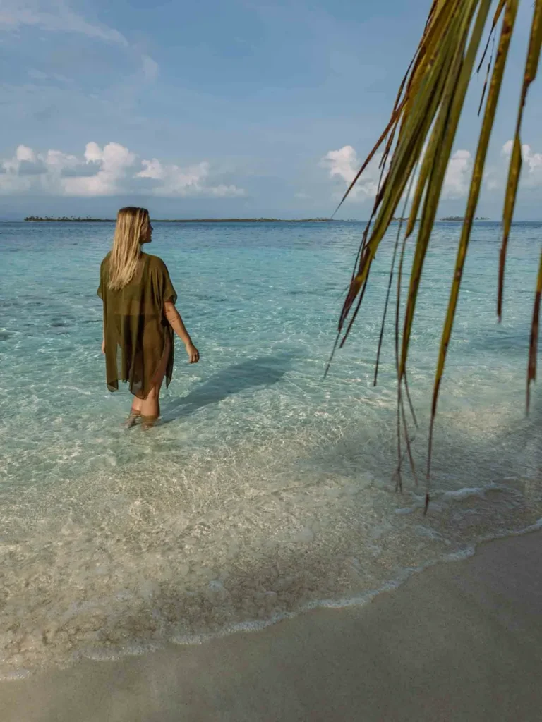 woman in a bikini with a green thing bathrobe walking through the shallow clear water on a sandy beach with some palm leaves hanging into the picture