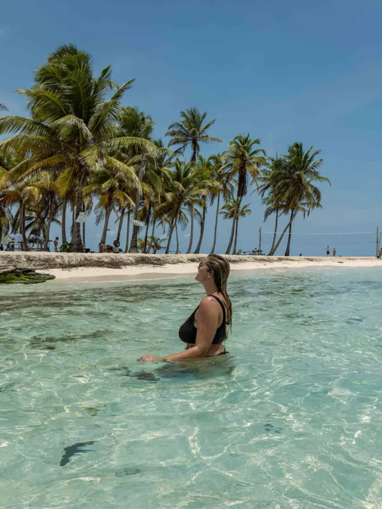 blonde woman with a black bikini and sunglasses kneeing in the shallow clear water at San Blas Islands - cant be missed on your ultimate panama itinerary, with white sand beaches, a volleyball net, tall palm trees and a few amenities in panama
