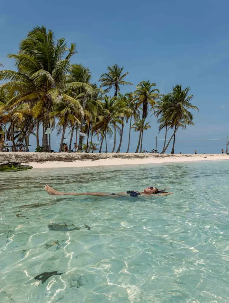 woman floating in the crystal clear ocean in front of a sandy island with coconut palmtrees
