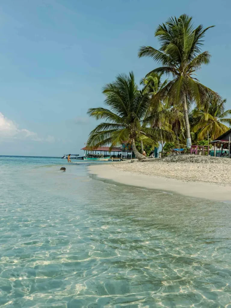 crystal clear shallow water at San Blas islands in panama with white sand palm trees and a few huts with amenities and a few boat in the back 