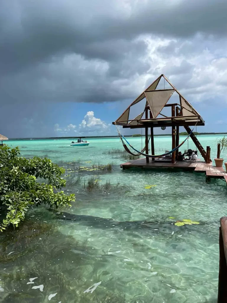 sea view with some plants in and around the water and a wooden gateway with a small bungalow with some hammocks and a boat in the back with some dark clouds on the sky in bacalar mexico