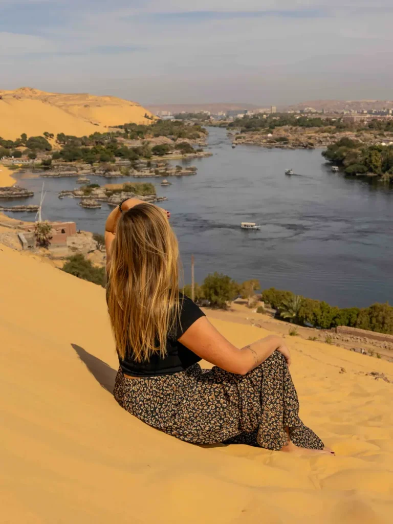 woman sitting on a sand dune with views of a lush river with palm trees in the distance