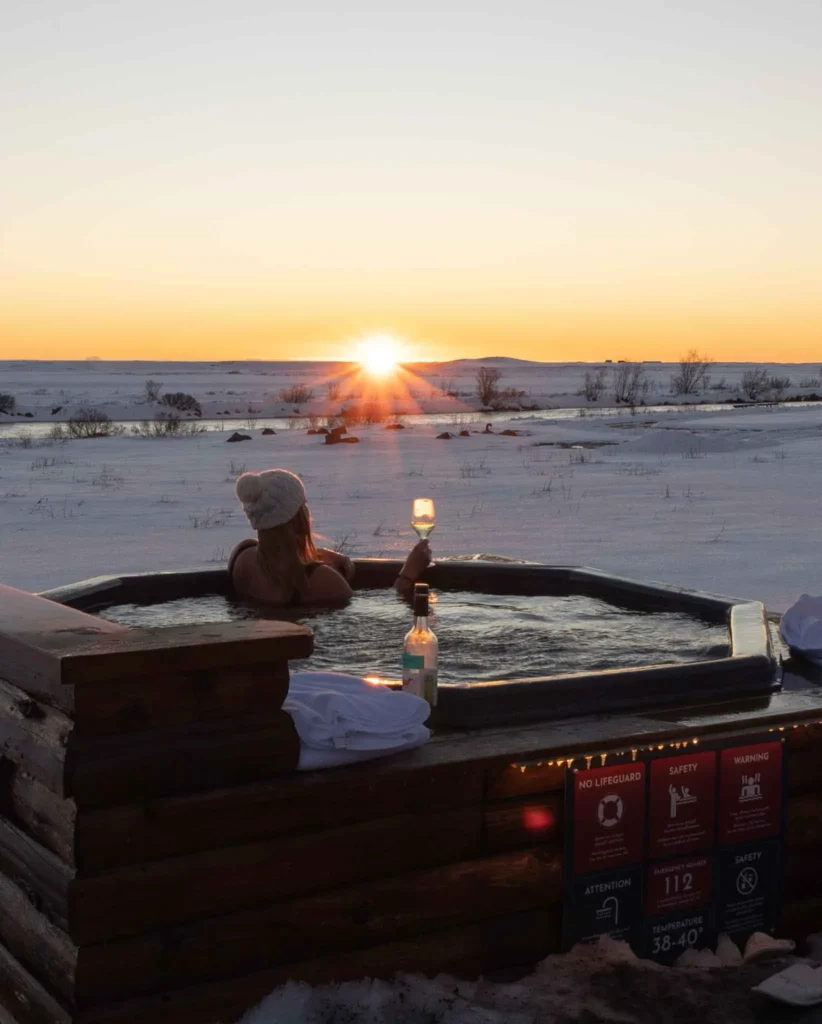 woman with a knitted hat in a wooden hot tub having a drink surrounded by snowy landscape at hotel ranga in iceland