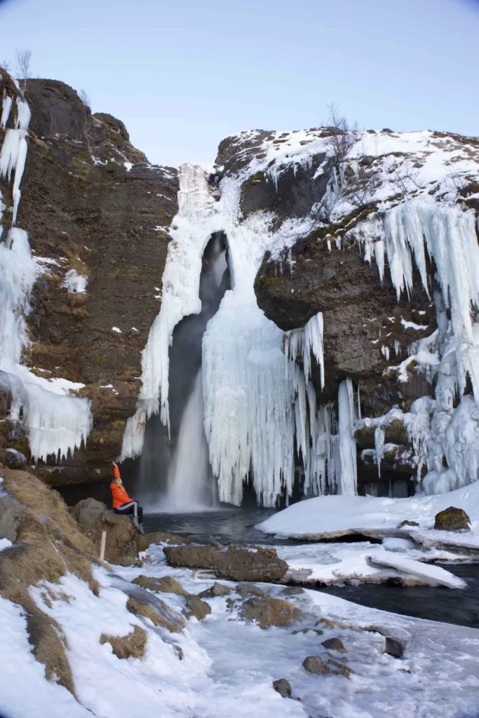 blonde woman with an orange winter jacket sitting in front of the frozen waterfall gluggafoss in volcanic rock in iceland