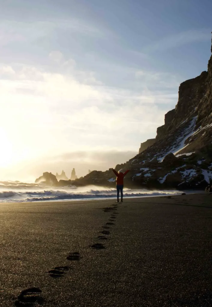 Vik Black Sand Beach woman stretching her arms on a black sand beach with big black rocks with a low sun at vik beach in iceland