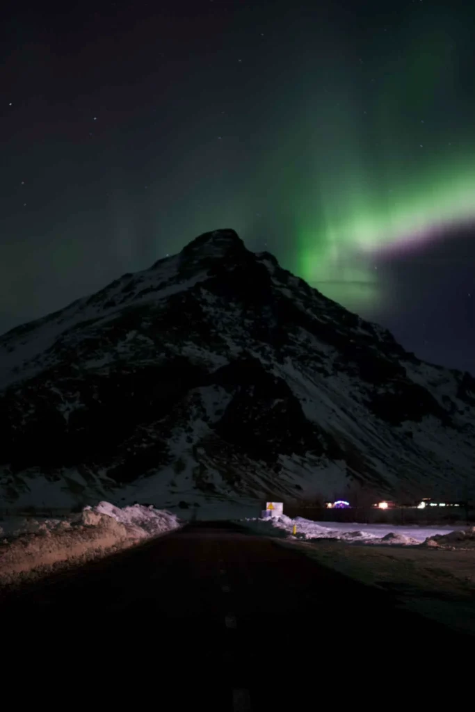 green northern lights visible behind a snowy mountain with a few small huts in the front
