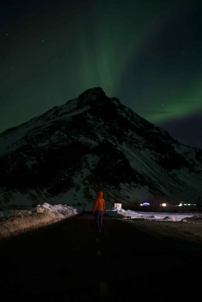person in an orange jacket standing in front of a mountain watching green northern lights with a few small huts in the back
