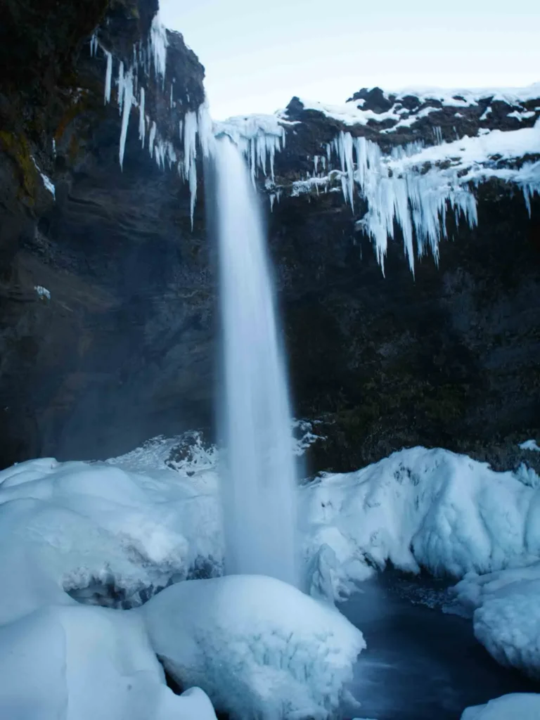 hidden gems in Iceland: Kvernufoss waterfall falling from black rocky terrain in a snowy landscape with some ice spikes at hidden gem waterfall kvernufoss in iceland