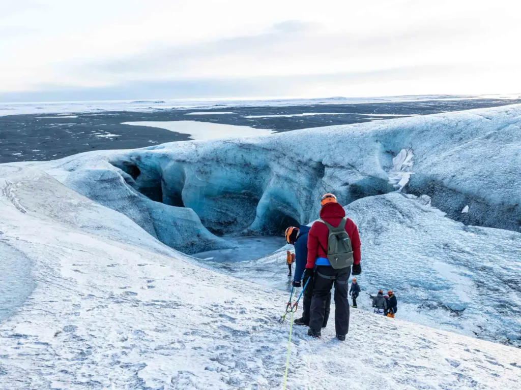 people in winter active outfits hiking a glacier that looks like a canyon made of ice, in the back is some black rock or sand landscape at Vatnajökull glacier in iceland