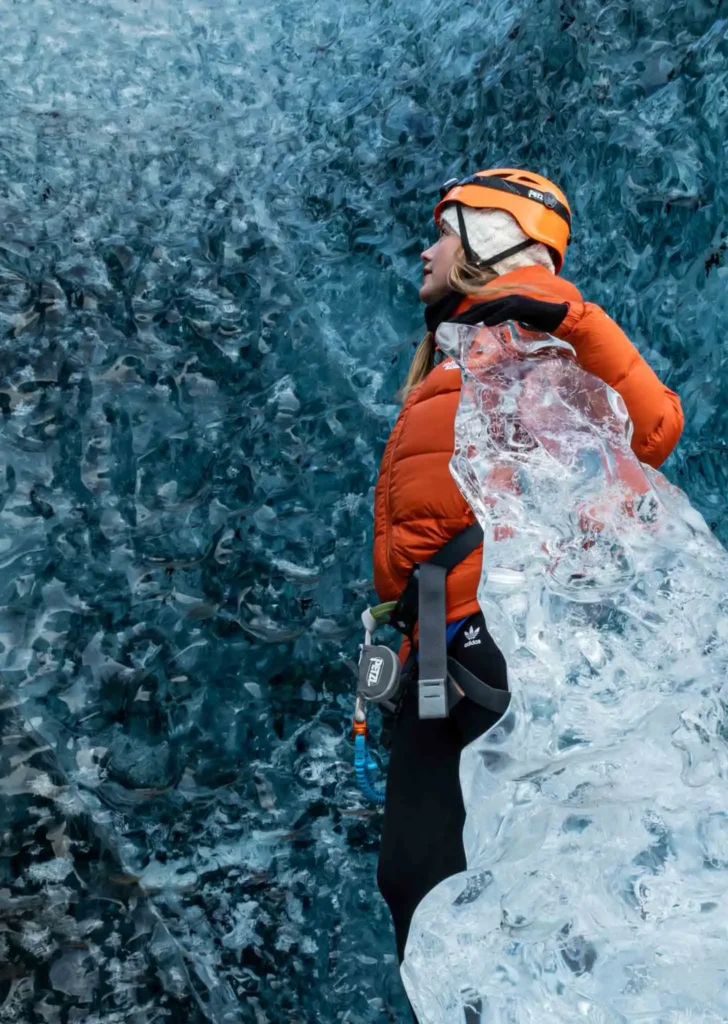woman in an orange winter jacket and an orange helmet starring into an ice wall with one icy peak in the front at the Vatnajökull glacier hike in iceland