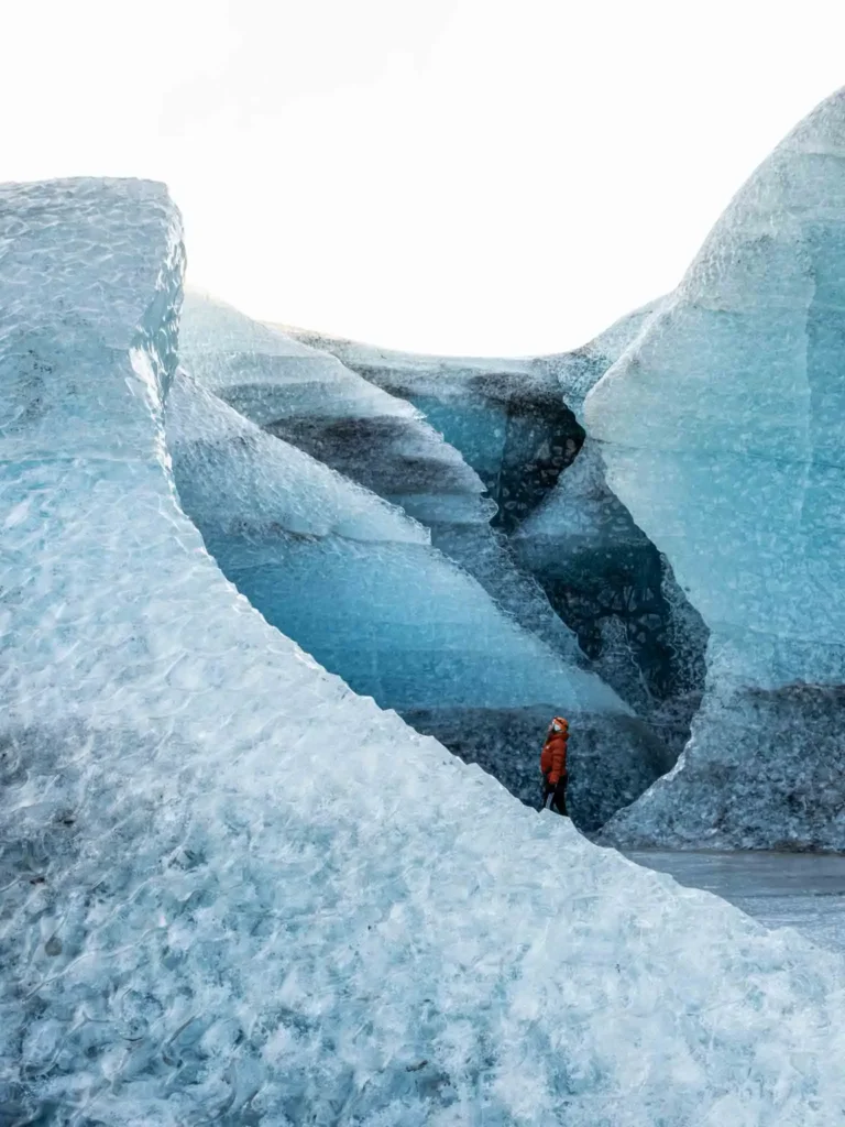 woman in orange winter jacket inbetween an ice canyon in iceland