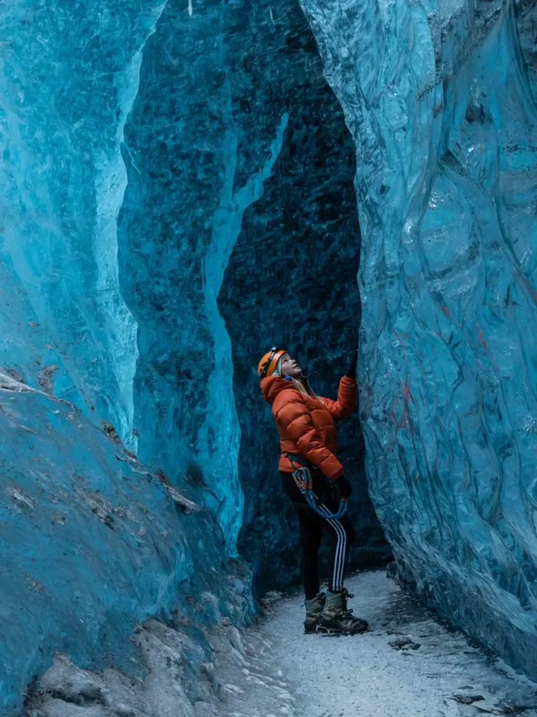 woman in an orange winter jacket standing inside the Vatnajökull glacier with blue ice in iceland