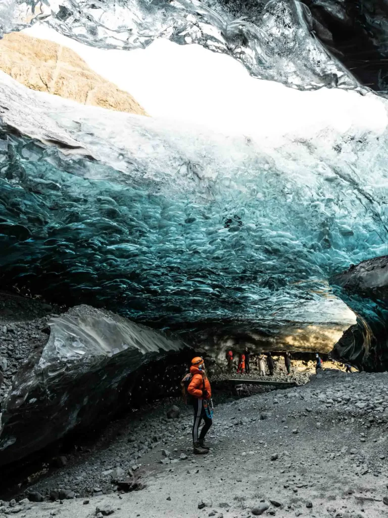 woman with an orange winter jacket and orange helmet starring at an ice wall inside a cave in iceland