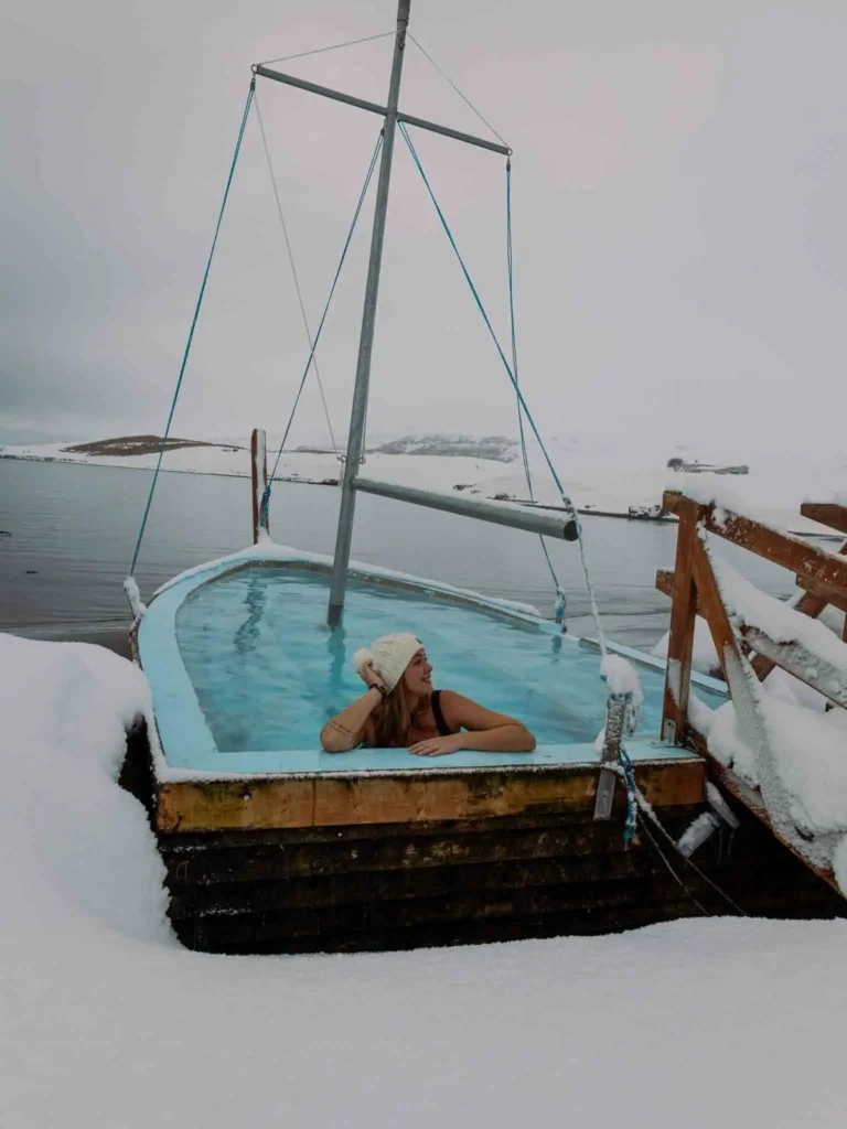 Hauganes hot tubs – one of my favorite hidden gems in Iceland woman with a white knitted hat in a boat, which is used as a pool, which appears to be in the sea with small snowy land areas visible in the back, there is also a small wooden gateway to the pool inside the boat at huaganes hot tubs in iceland