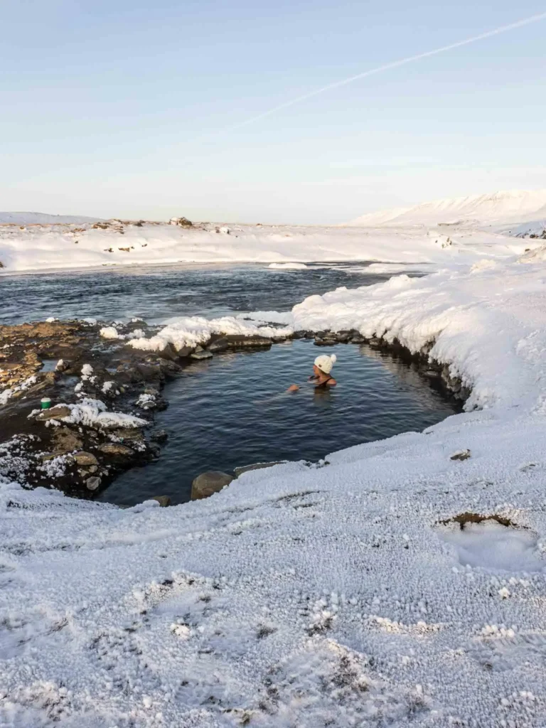 woman with a white knitted winter hat laying in a hot spring surrounded by a black rocky but snowy landscape at fosslaug hot spring in iceland
