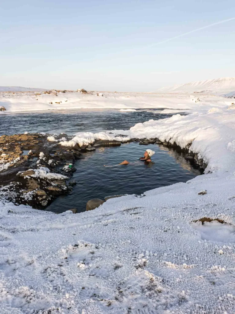 one of the best hidden gems in Iceland: Fosslaug hot spring woman with a white knitted winter hat laying in a hot spring surrounded by a black rocky but snowy landscape at hidden gem fosslaug hot spring in iceland