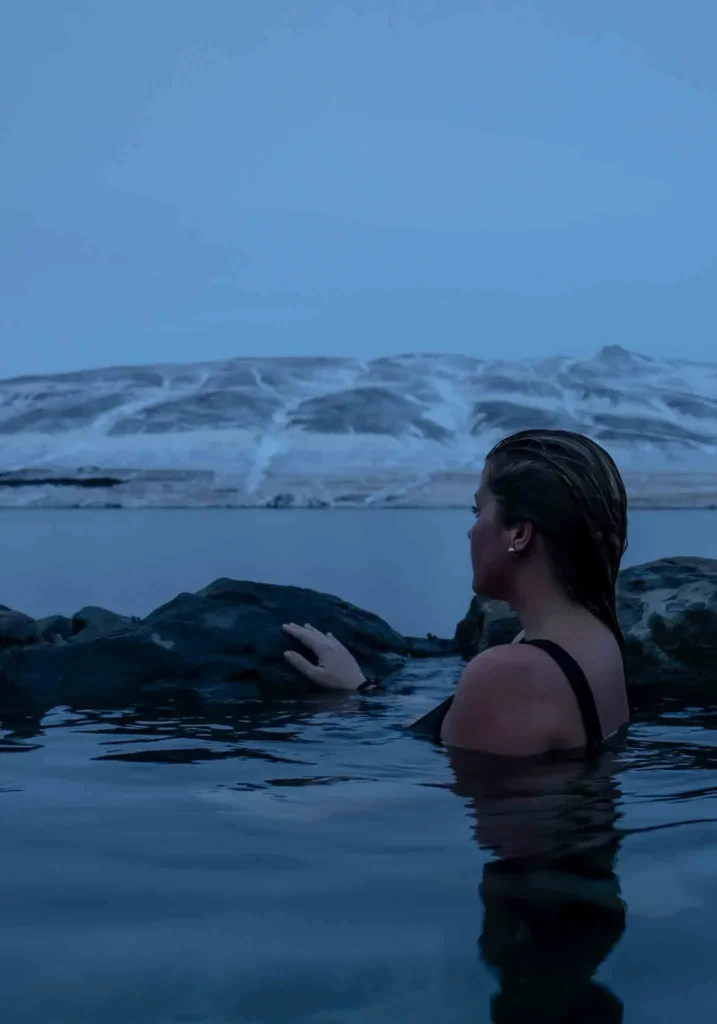 woman in a hot spring surrounded by black rocks and snowy hills and the sea in the background at hidden gem hvammsvik in iceland