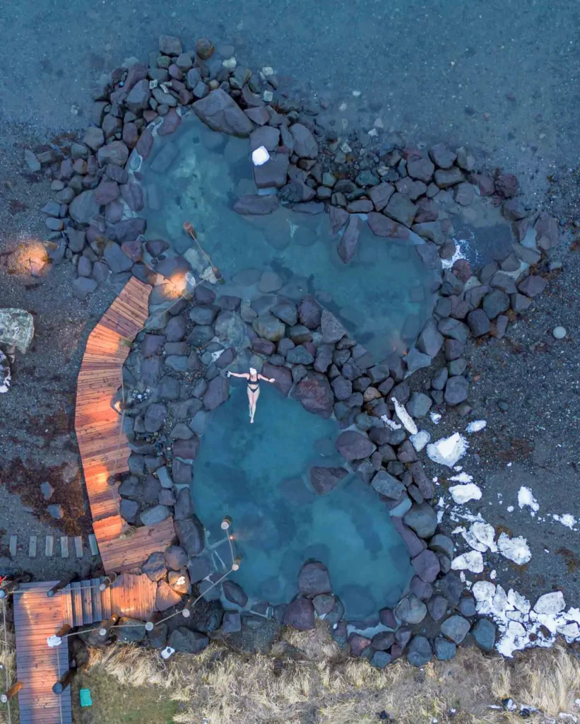 hidden gems in Iceland: Hvammsvik Nature Baths drone shot of a woman in a natural pool made by black volcanic rock at hvammsvik in iceland