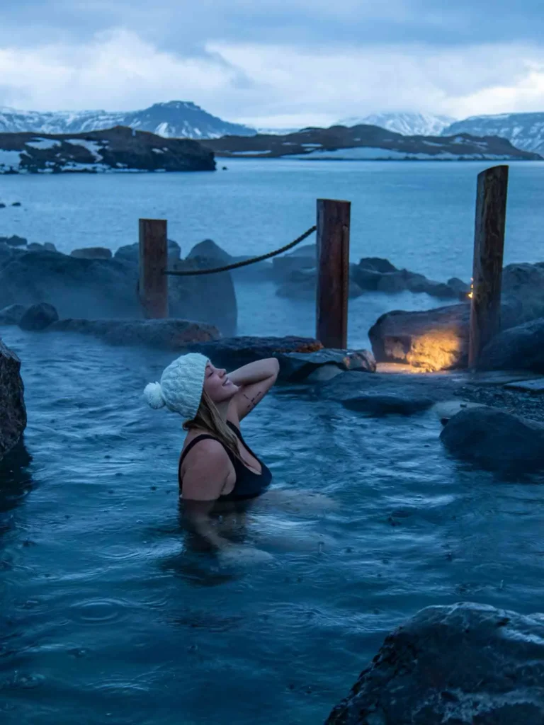 you want to pack your bikini for your trip to Iceland in winter woman wearing a white knitted hat looking at the sky while in a hot sping surrounded by black rocks. wooden poles, the sea and some partially snowy mountains in the back at hvammsvik in iceland