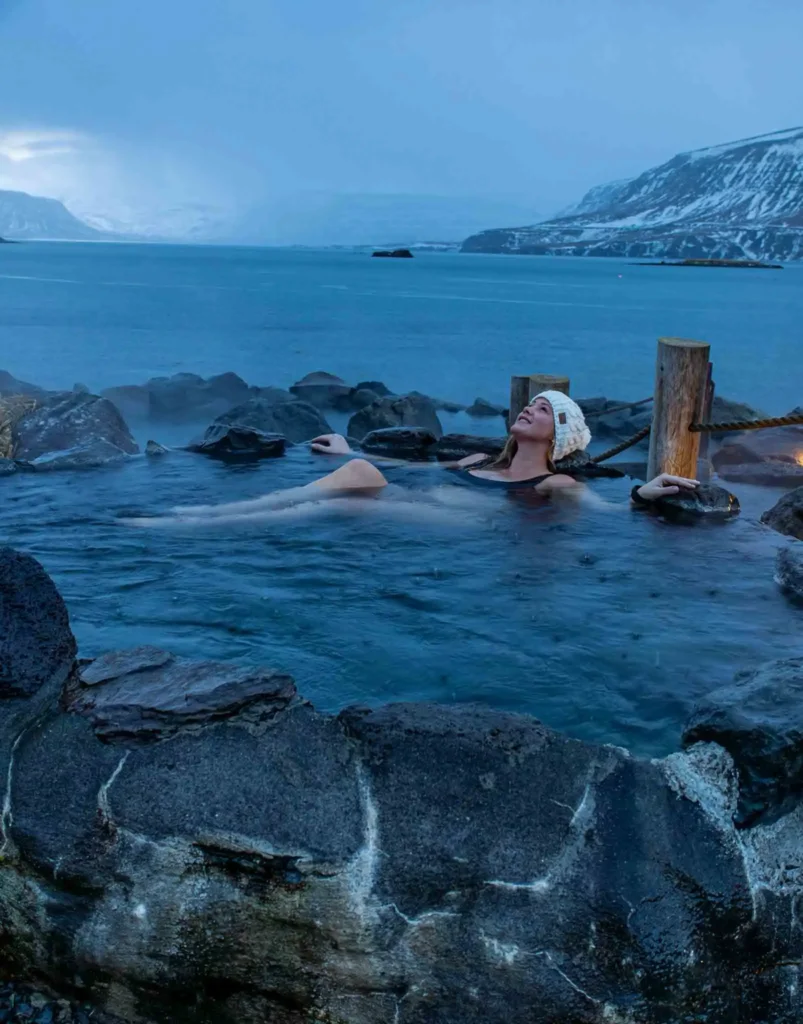 woman with a white knitted hat laying in a natural pool formed by dark rocks with a view on the sea and partially snowy hills in the back at hvammsvik nature baths in iceland