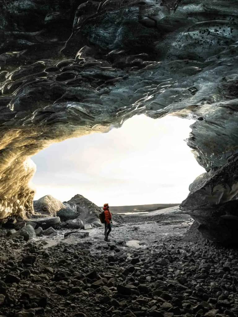 a cave within a dark landscape with rocks and sand, they cave has ice formation on its walls in iceland