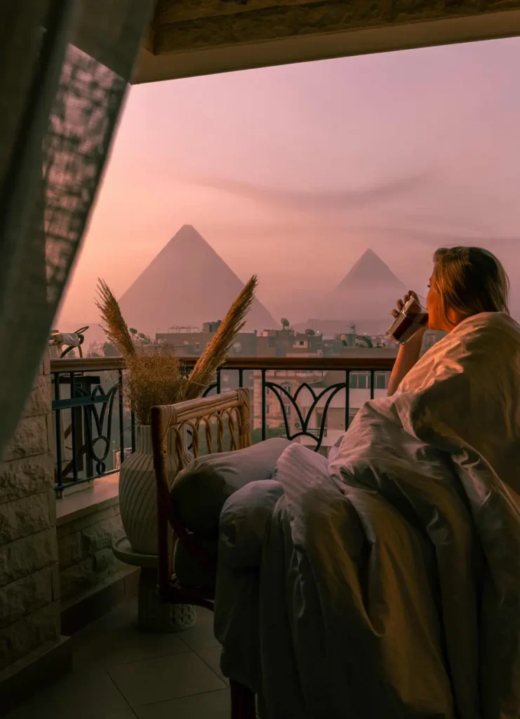 woman sitting on the balcony with a cup of coffee in her hand during sunrise, looking at the pyramids in the distance