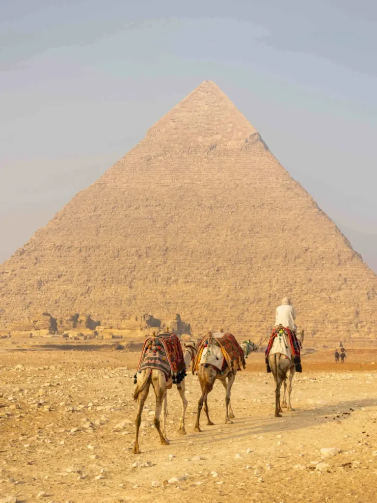 camels being ridden in the middle of the desert with a pyramid in the background