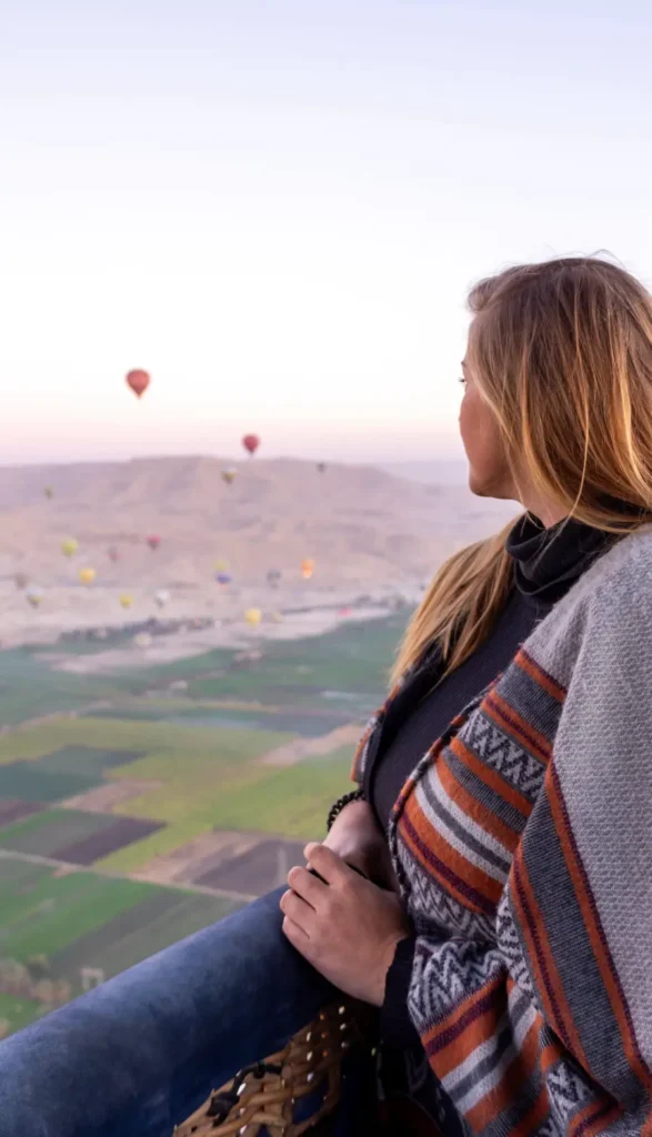 woman standing in a hot air balloon looking into the distance with more hot air balloons, mountains and blue skies during sunrise
