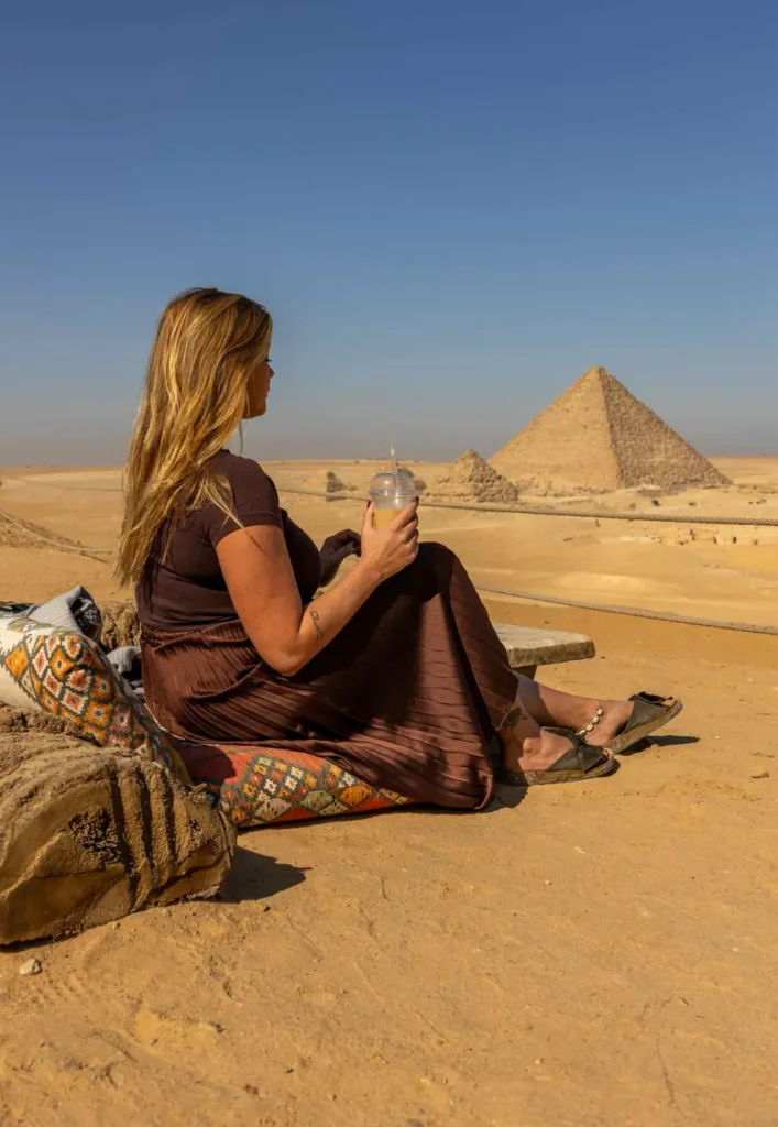 a woman sitting on a carpet in the desert with an iced coffee in her hand and view of the pyramids in the distance