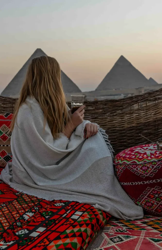 woman sitting on a rooftop terrace with colourful carpets looking at the pyramids in the distance