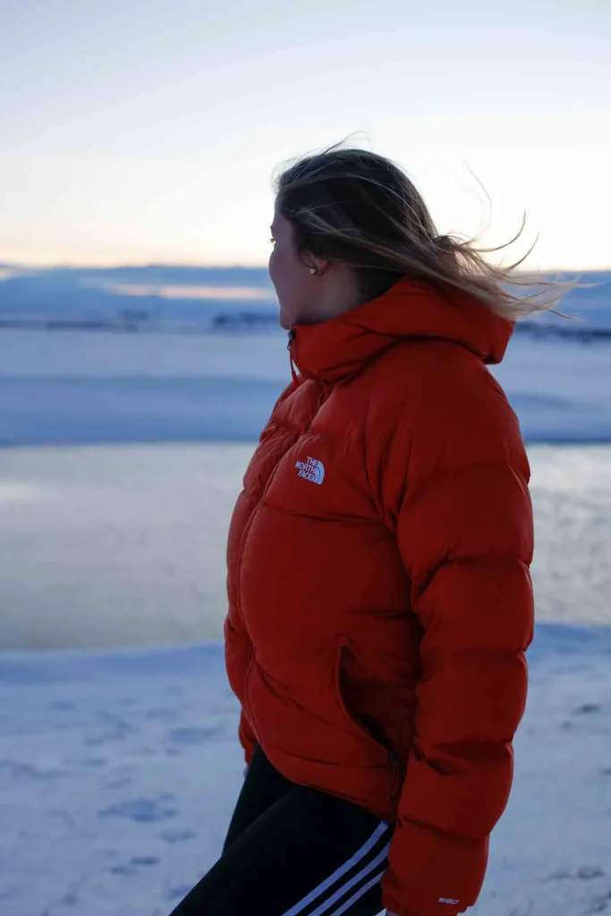 the north face keeping me warm during the cold winter days woman in an orange winter jacket looking at a snowy landscape in iceland