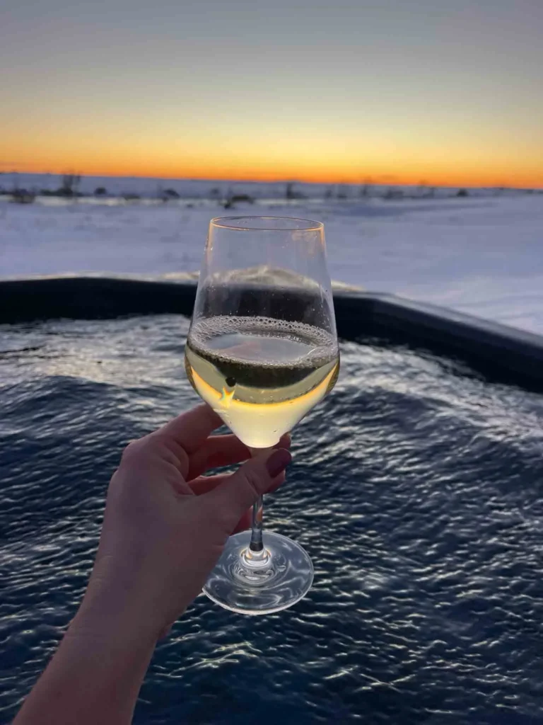glass of wine with a view person holding a glass of wine for a picture with a view of a hot tub and a snoey landscape at sunset or sunrise in iceland
