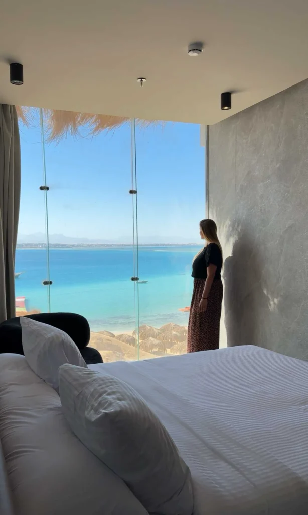 woman looking out onto the ocean from a hotel room with a large glass fron, curtains and king size bed with white sheets