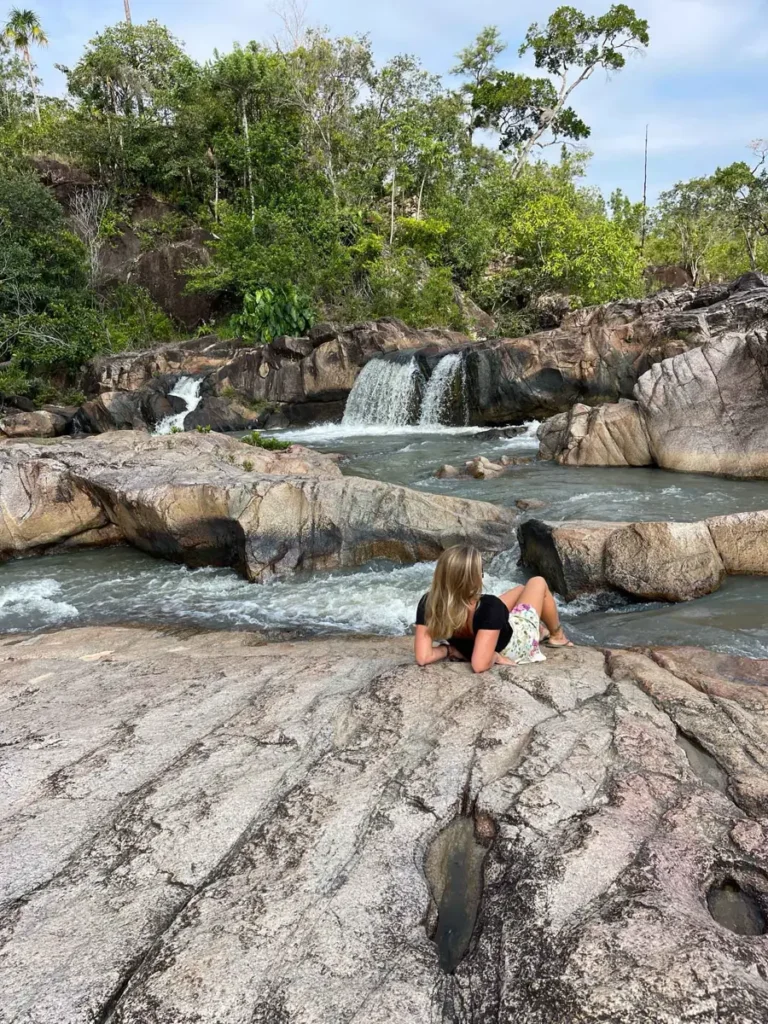 blonde woman laying on some big rocks forming multiple small pools and a small waterfall in the back surrounded by rocks and a pine forest at mountain pine ridge reserve in belize