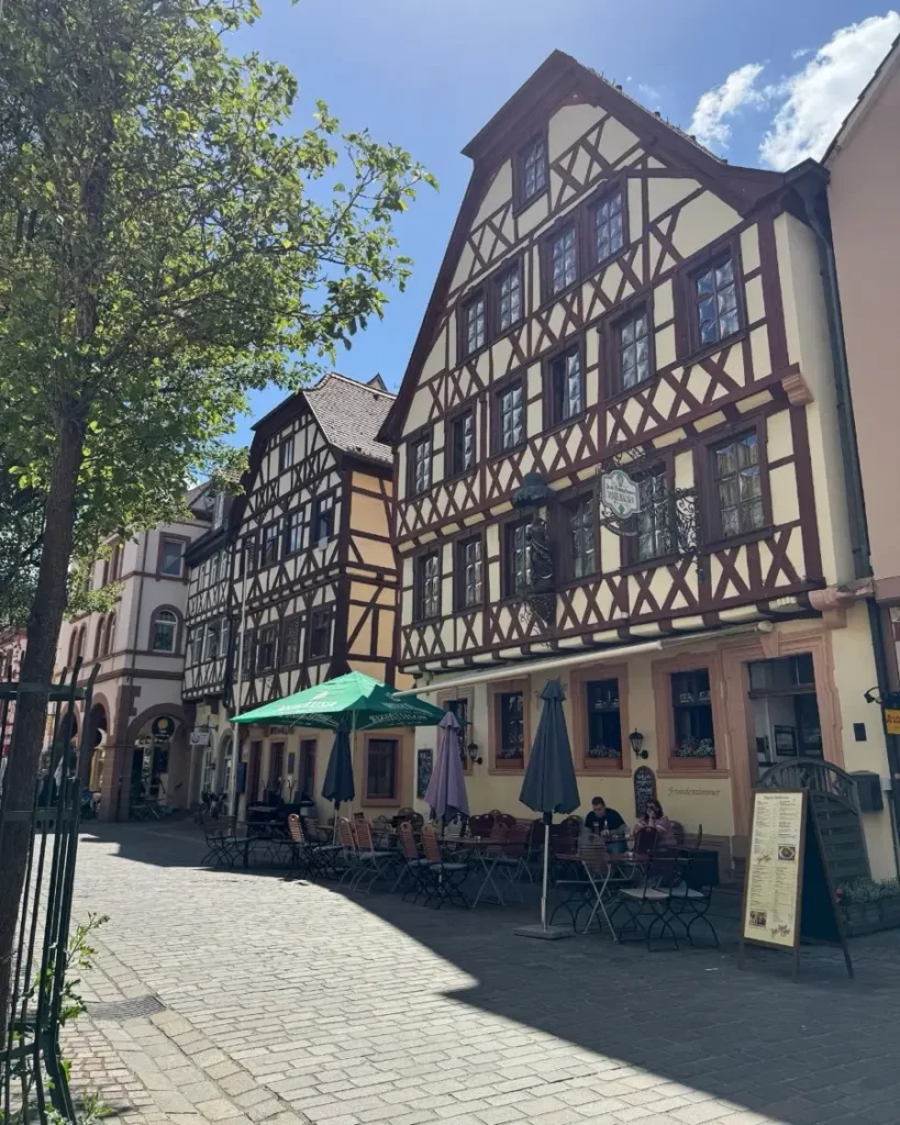 Traditional half-timbered houses on a cobblestone road with restaurant outdoor seating areas and some trees in Karlstadt, Germany