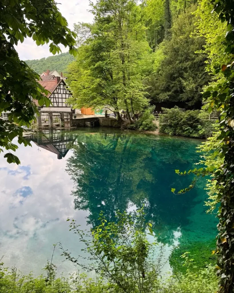 Mystical karst spring with clear blue water surrounded by green trees and one half-timbered traditional mill in Blaubeuren, Germany