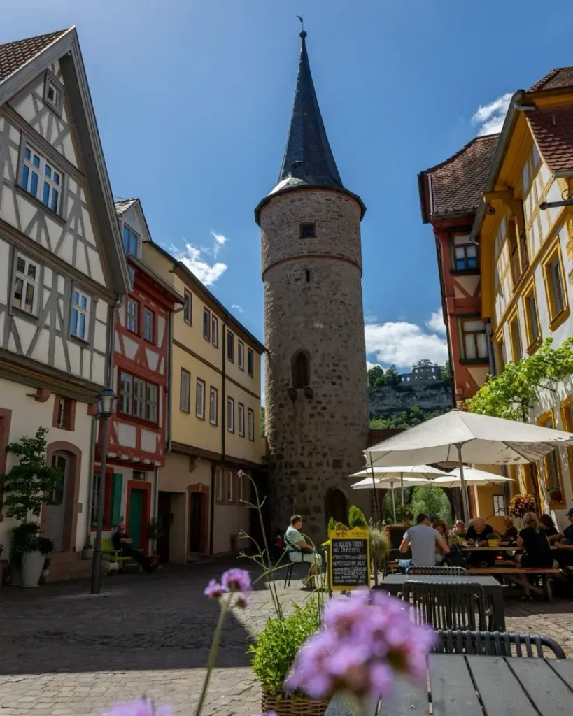 Old town with a historic watch tower with sharp roof, half-timbered houses, flowers and an outdoor seating are of a restaurant in Karlstadt, Germany