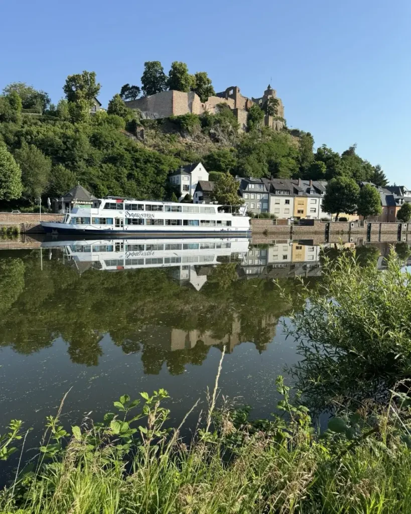Castle on a hill with trees and some houses around and a river ship docked in front in the fairytale town of Saarburg in Germany