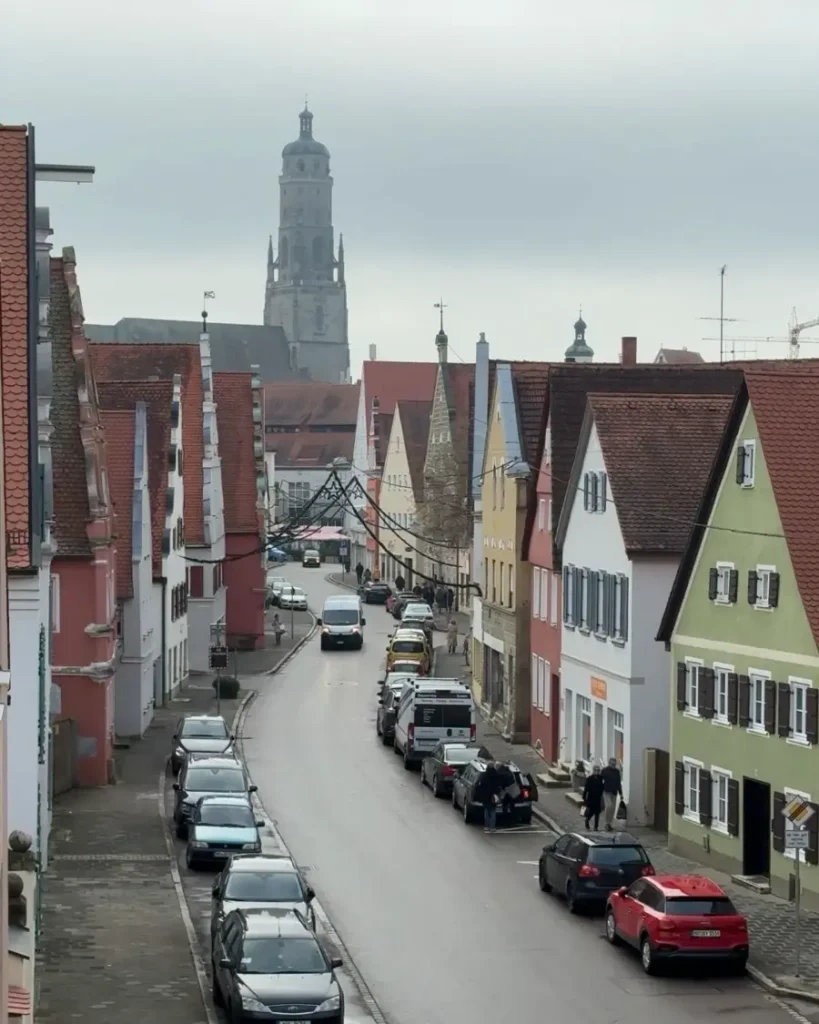 Road leading to a large church with colourful houses on each side and cars parked along the road in Nördlingen
