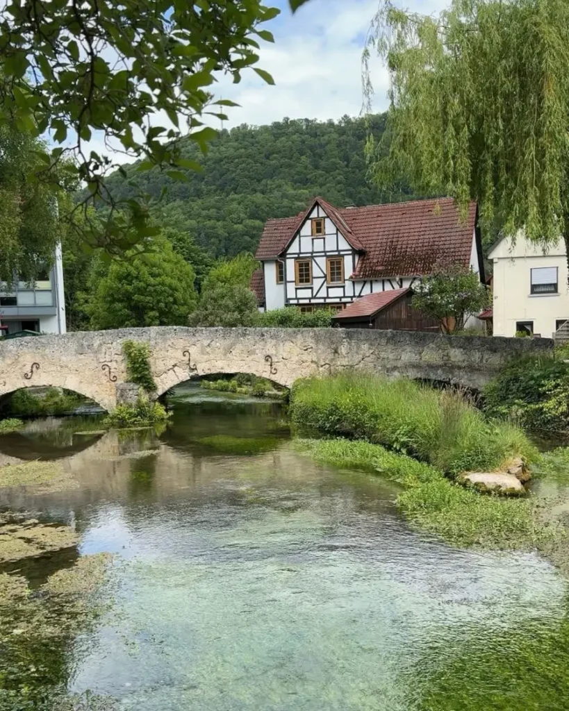 Historic bridge with multiple arches going over a small stream with water plants, surrounded by greenery and a half-timbered house in Lichtenstein, Germany