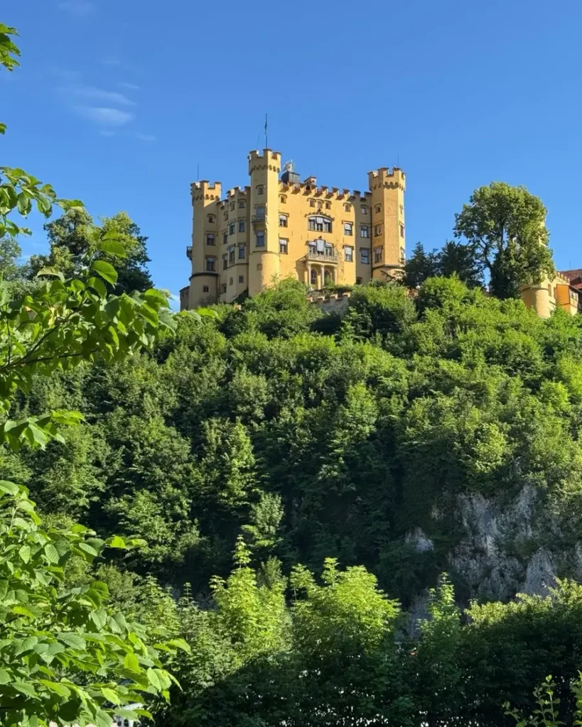 Yellow hillside castle with multiple towers on top of hill with green vegetation around - Hohenschwangau Castle